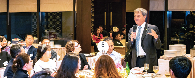 Alan Hasenfeld speaks while standing in front of several round banquet tables with seated people